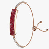 A rose gold bangle bracelet featuring a central curved bar paved with deep red crystals, accented with clear pavé stones at the top and bottom, and an adjustable sliding clasp on a light gray background.